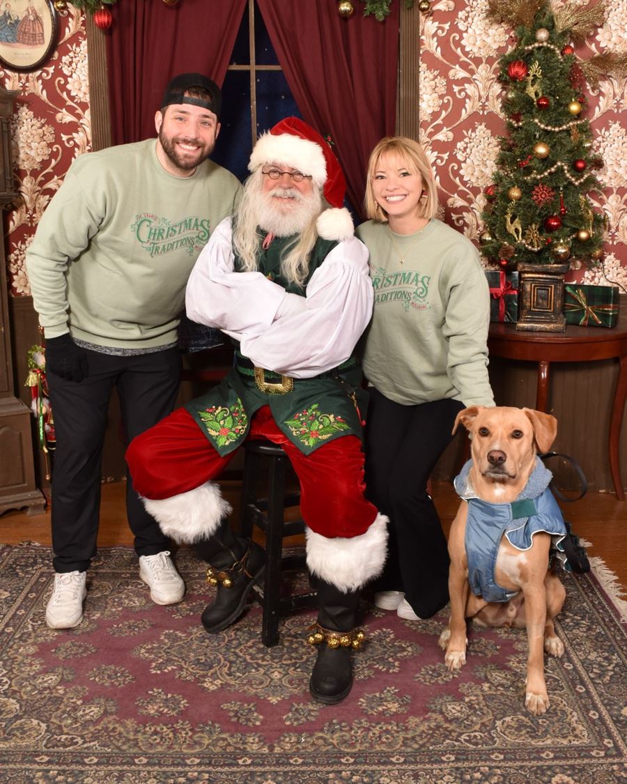A couple in Christmas Traditions sweatshirts with Santa and their dog in a blue coat
